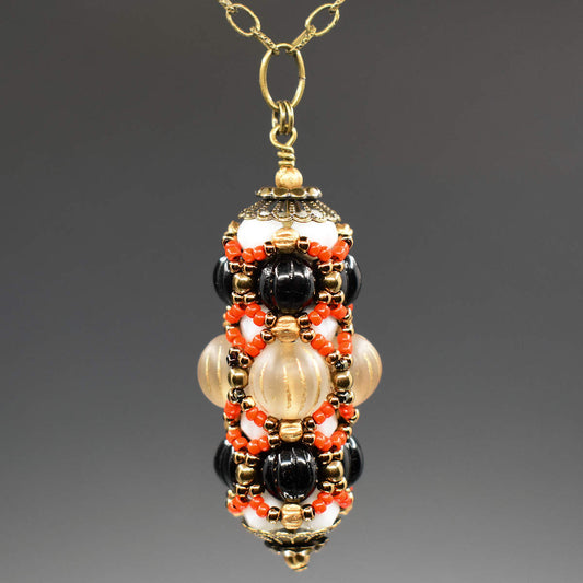 A black and cream pendant with coral accents hangs on a golden chain against a gray background. The pendant has a wider center row with frosted clear beads that have vertical gold stripes. The outer rows are both ridged black rounds. Smaller white beads helping form the structure are mostly hidden beneath the coral and gold seed bead netting that overlays the whole pendant. 