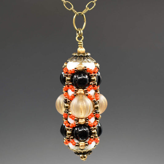 A black and cream pendant with coral accents hangs on a golden chain against a gray background. The pendant has a wider center row with frosted clear beads that have vertical gold stripes. The outer rows are both ridged black rounds. Smaller white beads helping form the structure are mostly hidden beneath the coral and gold seed bead netting that overlays the whole pendant.