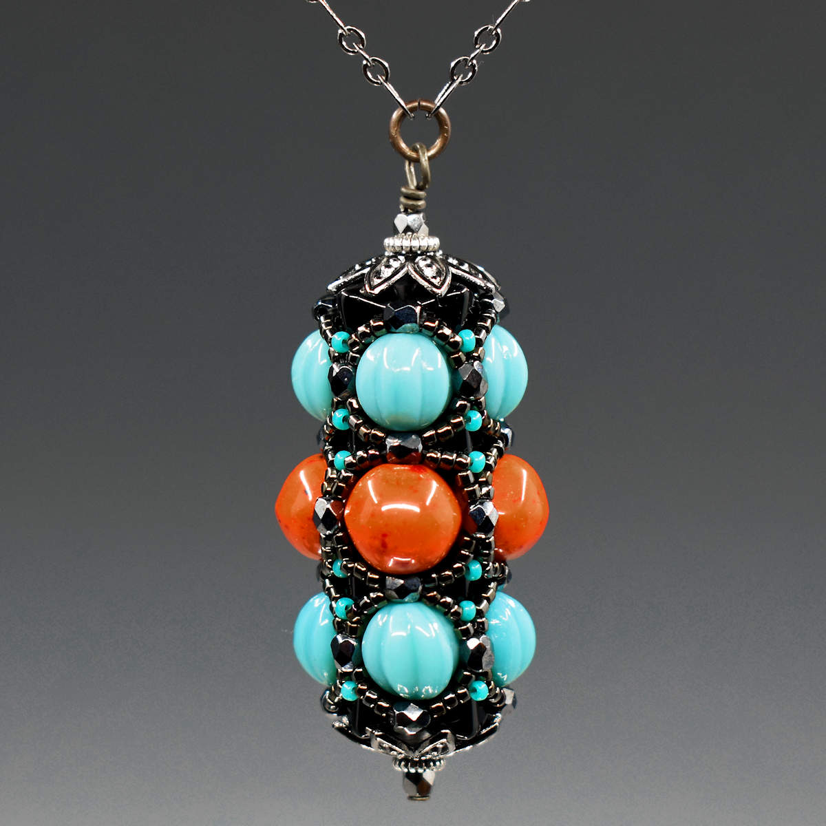 A turquoise and coral pendant on gunmetal chain hangs in front of a gray background. This pendant has turquoise outer rows and a coral red center row, all surrounded by a netting of black and gunmetal seed beads.