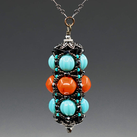 A turquoise and coral pendant on gunmetal chain hangs in front of a gray background. This pendant has turquoise outer rows and a coral red center row, all surrounded by a netting of black and gunmetal seed beads.