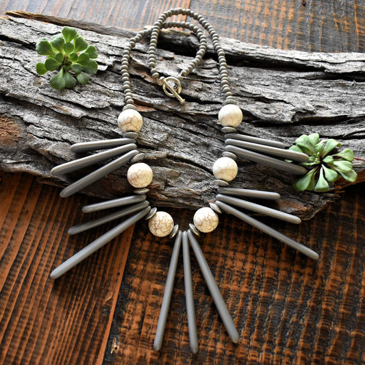 A necklace with long gray spine drops and soft cream rounds lays over a wood backdrop. The necklace has five sections of three gray spines, the outer ones grouped from shortest to longest towrds the center. The center section is formed with only the longest spines. Between the gray beads are soft cream round beads with light brown crackle lines on them. The back of the necklace is formed from smaller gray seed beads and a silver toggle clasp.