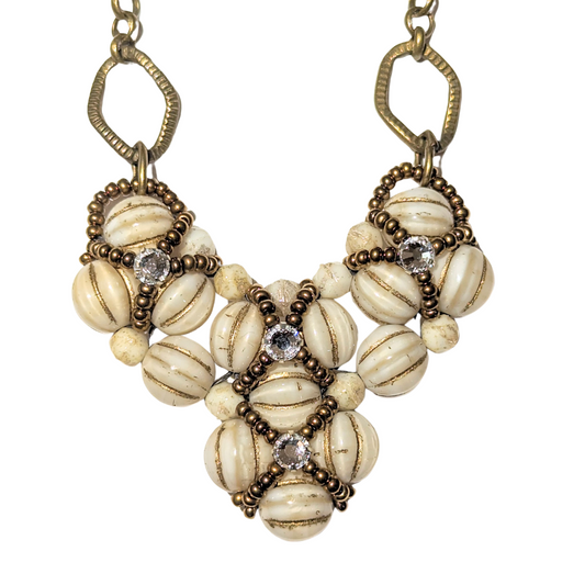 Cream and gold heart shaped pendant on brass chain. The heart is made from gold-striped cream beads overlaid with X-patterns of gold seed beads with clear rhinestones in the center.