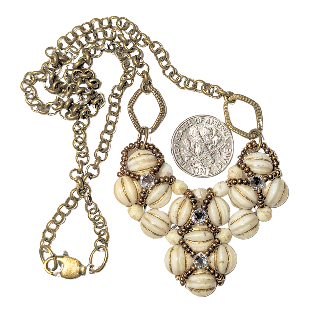 Cream and gold heart shaped pendant on brass chain, shown with a dime for scale. The heart is made from gold-striped cream beads overlaid with X-patterns of gold seed beads with clear rhinestones in the center.