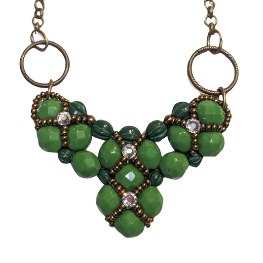 A wide green beaded heart pendant with brass chain on a white background. The heart is made from medium green beads with darker green melon beads on the edges. The beadwork has an overlay of gold seed bead X-shapes with clear rhinestones in their centers. 