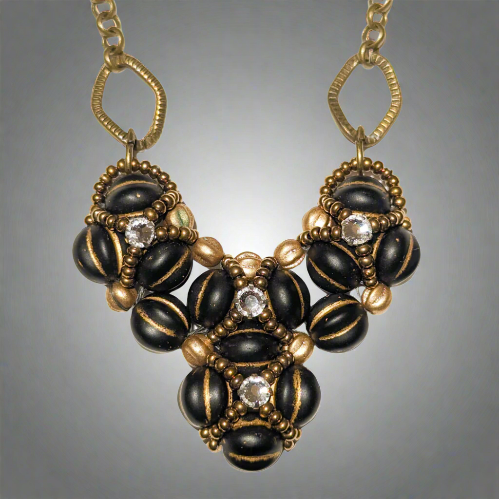 Heart shaped pendant on brass chain. The heart is made from gold-striped black beans overlaid with X-patterns of gold seed beads with clear rhinestones in the center.
