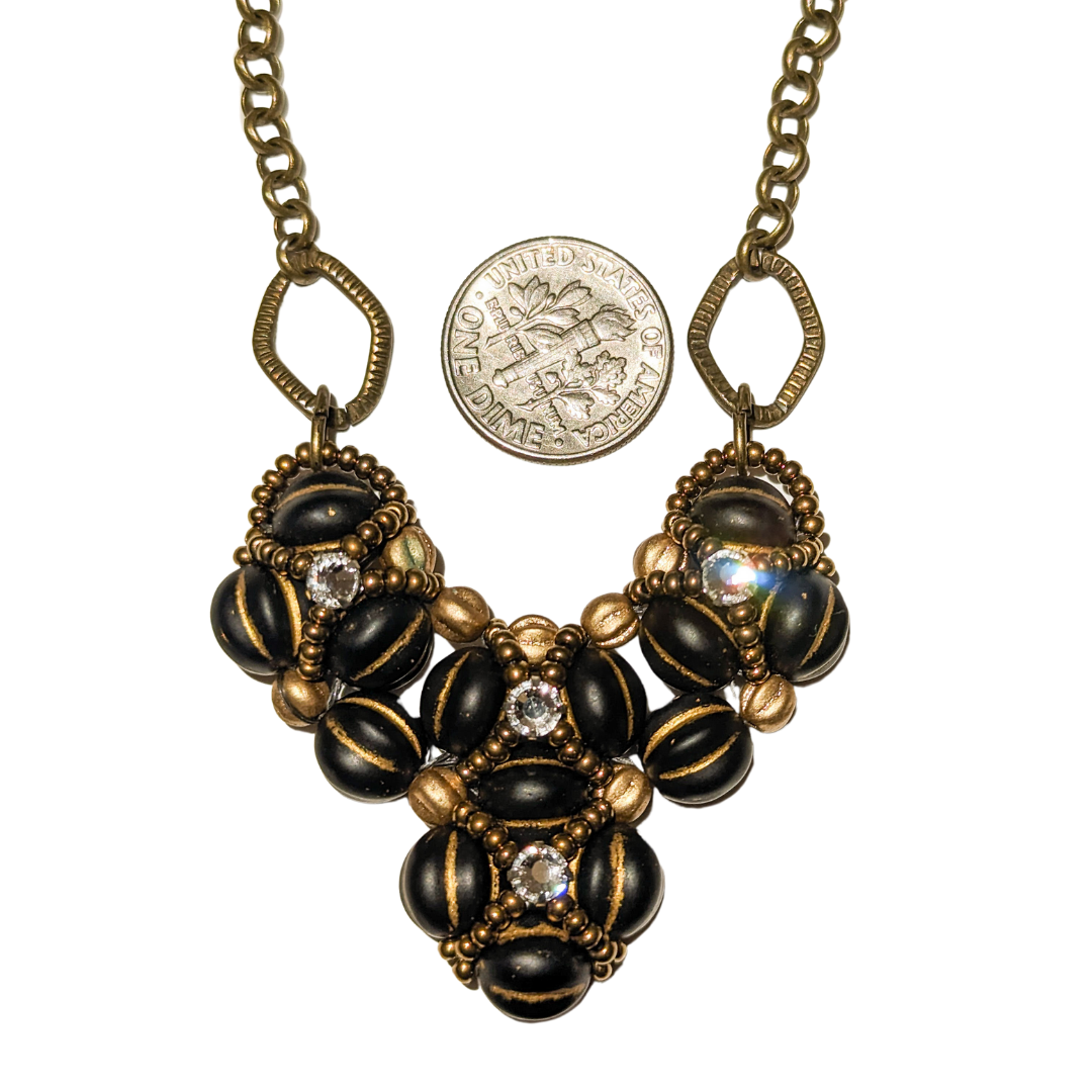 Heart shaped pendant on brass chain, shown with a dime for scale. The heart is made from gold-striped black beans overlaid with X-patterns of gold seed beads with clear rhinestones in the center.