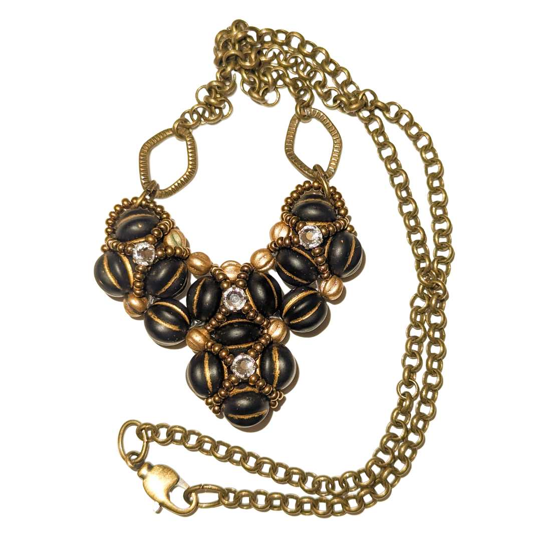 Heart shaped pendant on a brass chain with lobster claw clasp. The heart is made from gold-striped black beans overlaid with X-patterns of gold seed beads with clear rhinestones in the center.