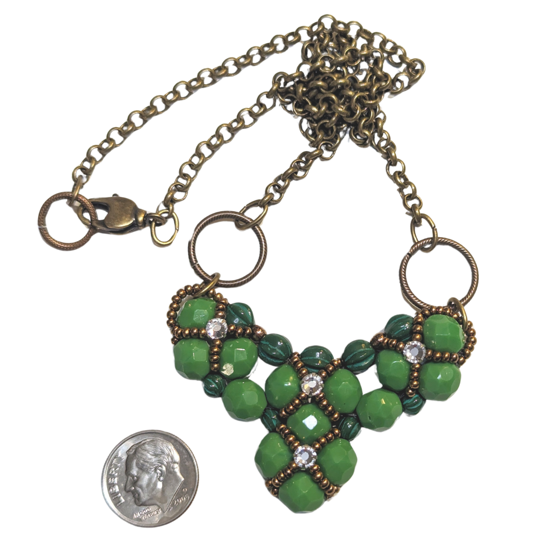 A wide green beaded heart pendant with brass chain on a white background. The heart is made from medium green beads with darker green melon beads on the edges. The beadwork has an overlay of gold seed bead X-shapes with clear rhinestones in their centers. A dime is shown for scale.