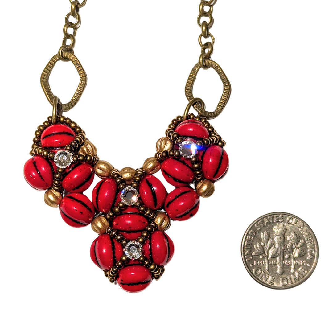 A red beaded heart pendant on a white background. The heart is made from red beads with black lines, with an overlay of gold seed bead X-shapes with clear rhinestones in their centers. A dime is shown for scale.