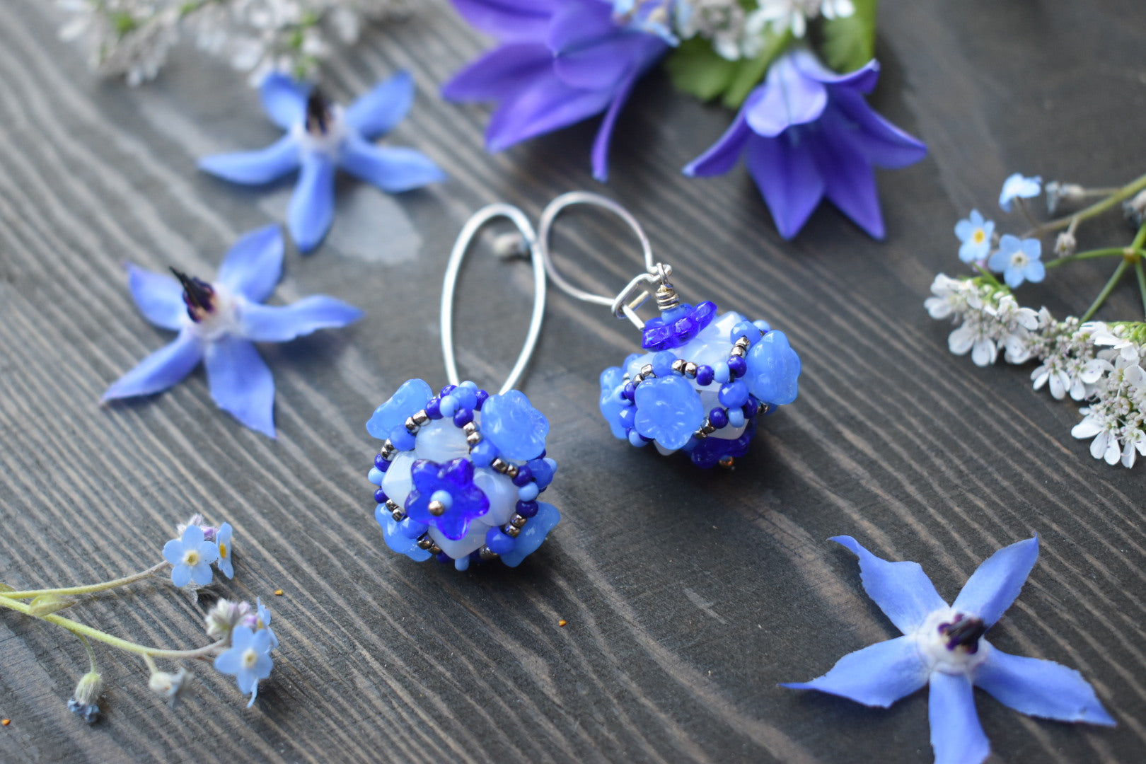 Light blue ball earrings with flowers on them lay on a wood background scattered with blue and white flowers.