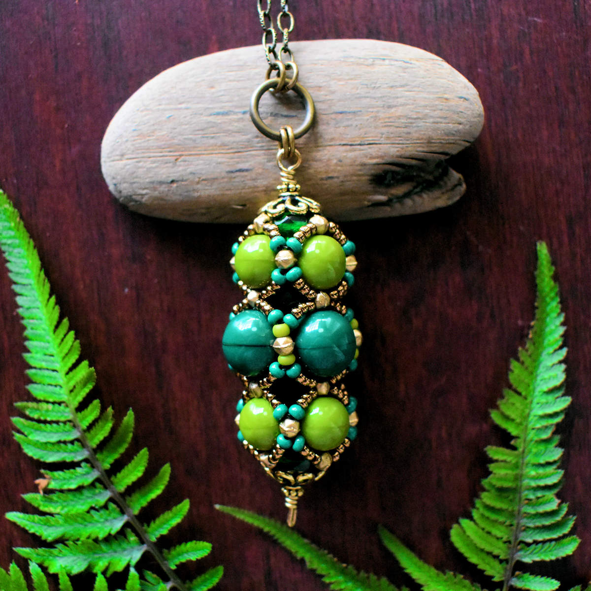 A green columnar pendant with three rows of round green beads outlined in gold seed beads lays against a small piece of driftwood on a red wood background. There are green ferns laying around the pendant.