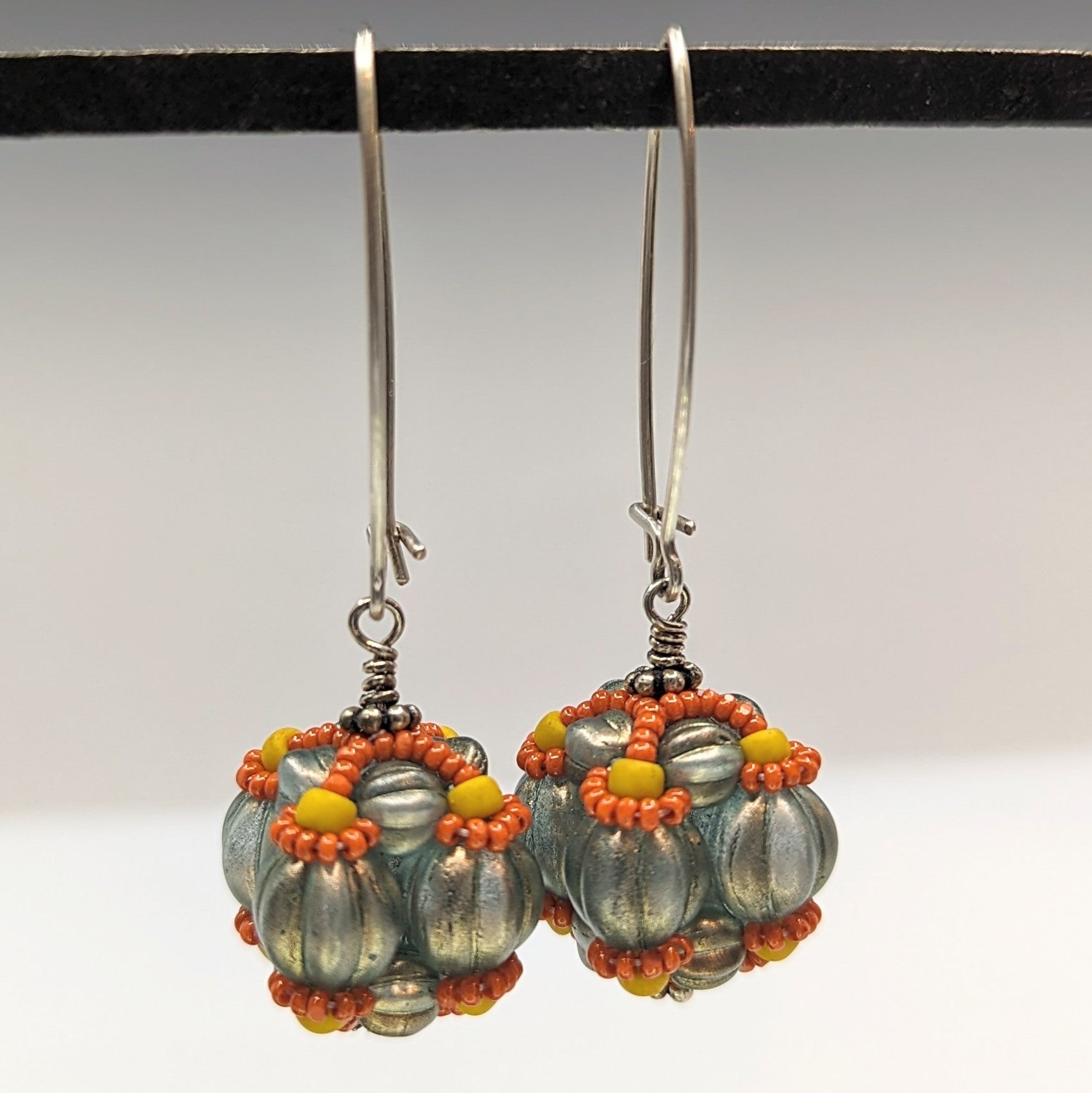 A pair of earrings hang from a black bar, against a gray background. The earrings have long silver oval wires that latch and there is a beaded ball dangling from the bottom that is made from ridged round beads that are a pale, translucent aqua color with gold finish. The top part of each beaded bead is outlined with lines of tiny yellow and orange-brown seed beads.