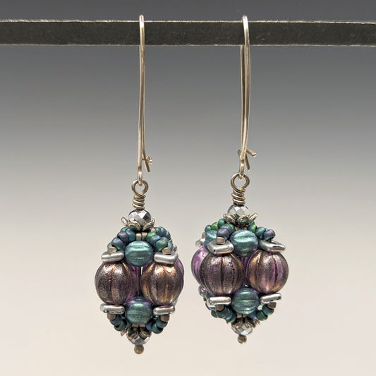 A pair of earrings hang from a black bar, against a gray background. The earrings have long silver oval wires that latch and there is a beaded ball dangling from the bottom that is made from ridged purple rounds surrounded by layers of silver and metallic green beads.