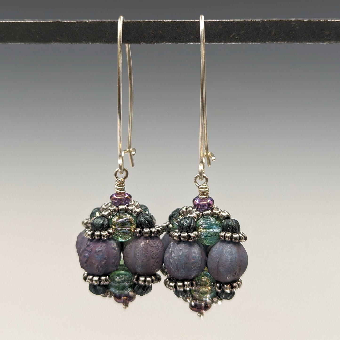 A pair of earrings hang from a black bar, against a gray background. The earrings have long silver oval wires that latch and there is a beaded ball dangling from the bottom of each earring. The beaded beads are s made from rough textured lilac beads surrounded by layers of silver and sea green beads. 