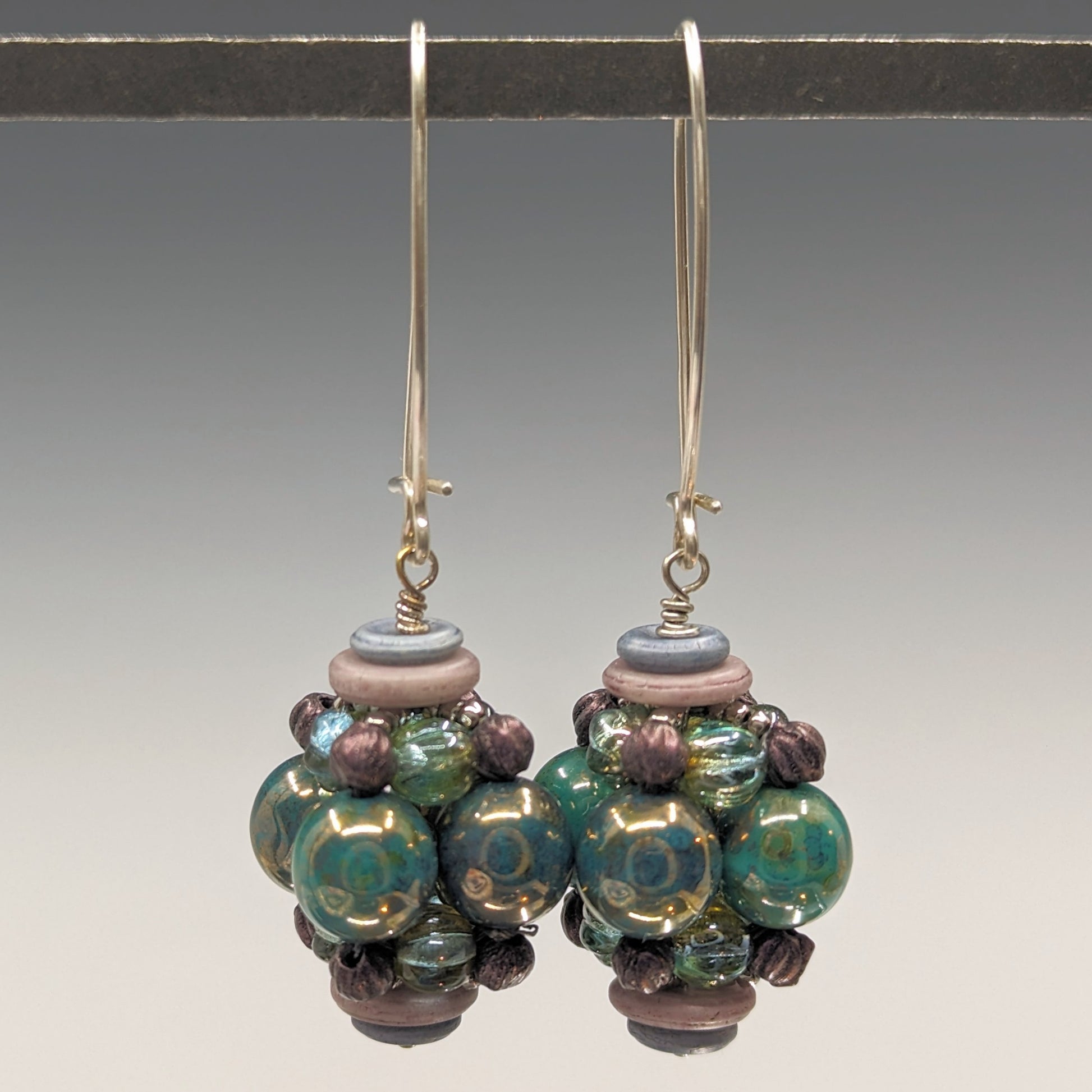 A pair of earrings hang from a black bar against a gray background. The earrings have long silver oval wires that latch and there is a beaded ball dangling from the bottom of each earring. The beaded beads are s made from bluish green beads with a bronze luster, surrounded by layers of silver, sea green, and lillac beads. 