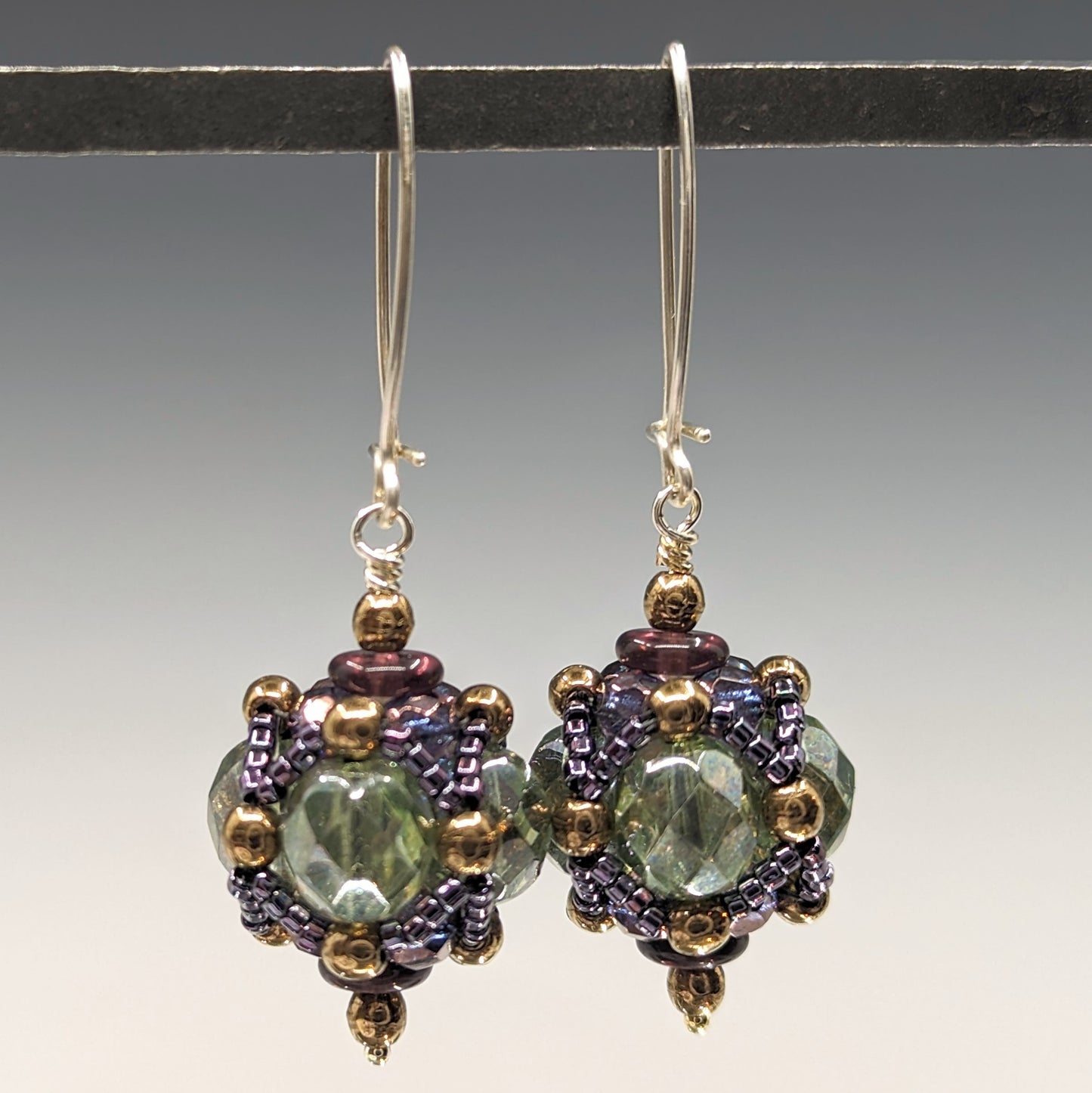 A pair of earrings hang from a black bar against a gray background. The earrings have long silver oval wires that latch and there is a beaded ball dangling from the bottom of each earring. The beaded beads are made from pale transparent green beads that have a layer of netted putple and bronze beads wrapped around them.