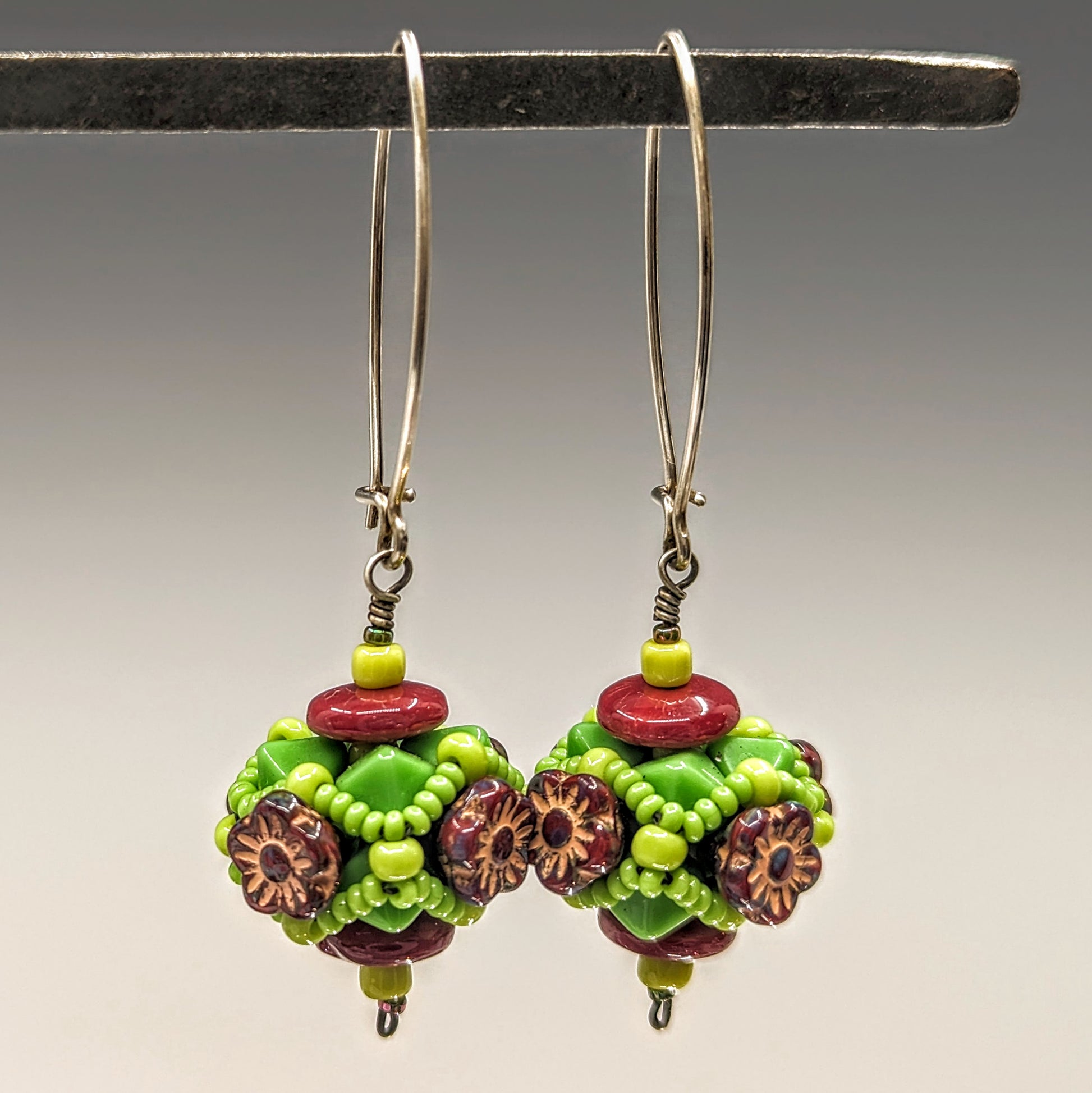 A pair of earrings hang from a black bar, against a gray background. The earrings have long silver oval wires that latch and there is a beaded ball dangling from the bottom that has burgundy flowers surrounded by a web of spring green seed beads.