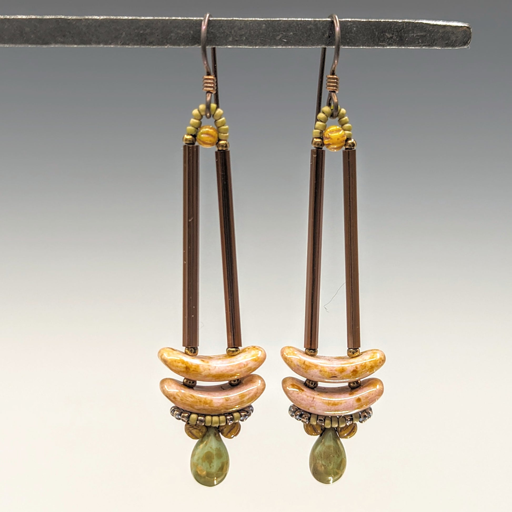 Earrings that look similar to a medicine dropper hang from  a black bar. There are two long, brown beads forming narrow vertical columns ending at the top of two speckled pink stacked arc shaped beads. At the bottom is a matte speckled turquoise green drop shaped bead, surrounded by a belt of matte gold seed beads.