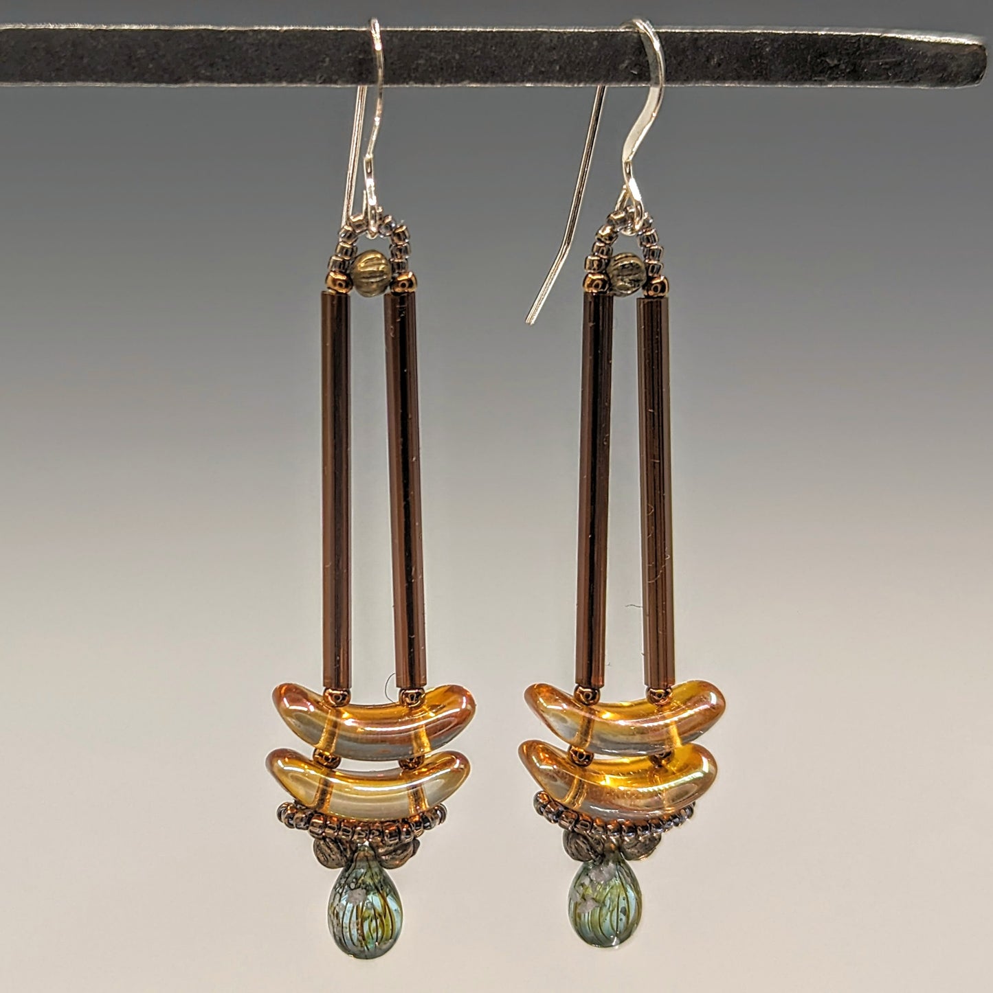 Earrings that look similar to a medicine dropper hang from  a black bar. There are two long, brown beads forming narrow vertical columns ending at the top of two transparent peach colored stacked arc shaped beads. At the bottom is a drop shaped bead made from aqua colored glass with black stripes inside of it, surrounded by a belt of gold seed beads.