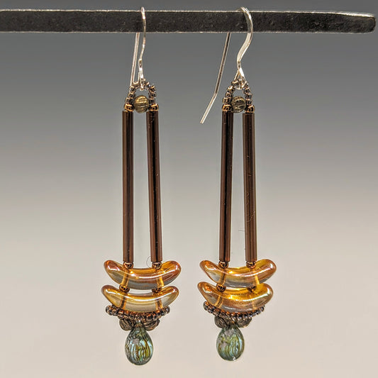 Earrings that look similar to a medicine dropper hang from  a black bar. There are two long, brown beads forming narrow vertical columns ending at the top of two transparent peach colored stacked arc shaped beads. At the bottom is a drop shaped bead made from aqua colored glass with black stripes inside of it, surrounded by a belt of gold seed beads.