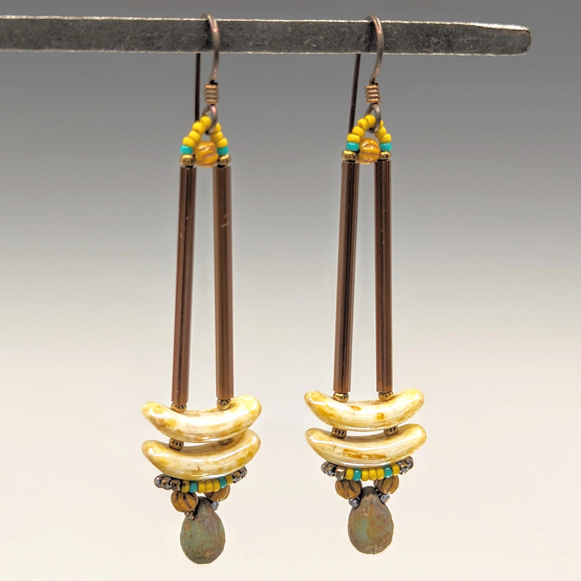 Earrings that look similar to a medicine dropper hang from  a black bar. There are two long, brown beads forming narrow vertical columns ending at the top of two speckled cream stacked arc shaped beads. At the bottom is a rough brown and turquoise drop , surrounded by a belt of mustard and turquoise seed beads.