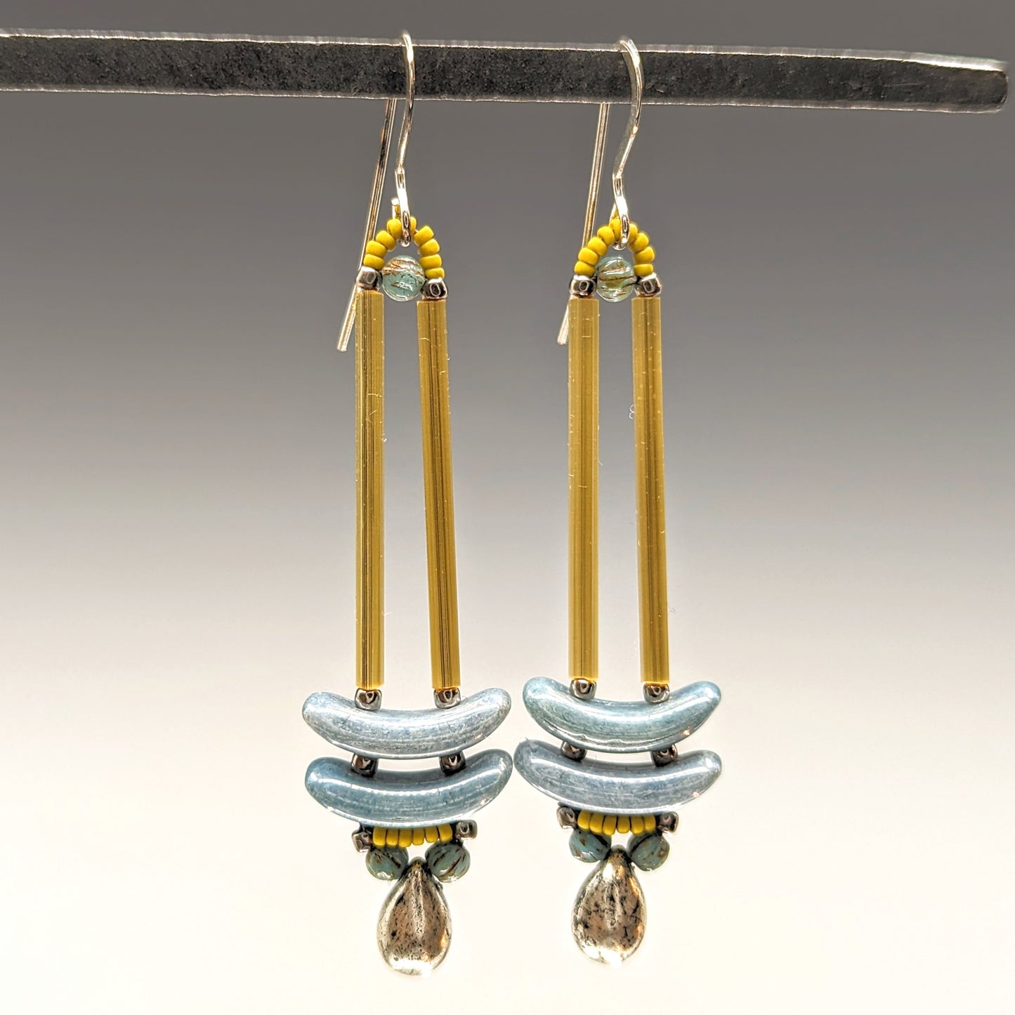 Earrings that look similar to a medicine dropper hang from a black bar. There are two long, yellow-gold beads forming narrow vertical columns ending at the top of two pale blue stacked arc shaped beads. At the bottom is a silver drop shaped bead between two turquoise rounds, all surrounded by a belt of mustard yellow seed beads.