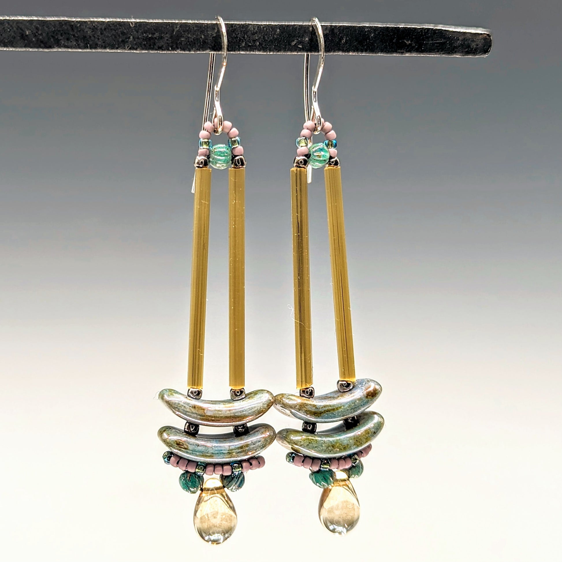 Earrings that look similar to a medicine dropper hang from a black bar. There are two long, yellow-gold beads forming narrow vertical columns ending at the top of two speckled gray-blue stacked arc shaped beads. At the bottom is a drop shaped bead made from clear champagne colored glass, surrounded by a belt of pale pink and dusty turquoise seed beads.
