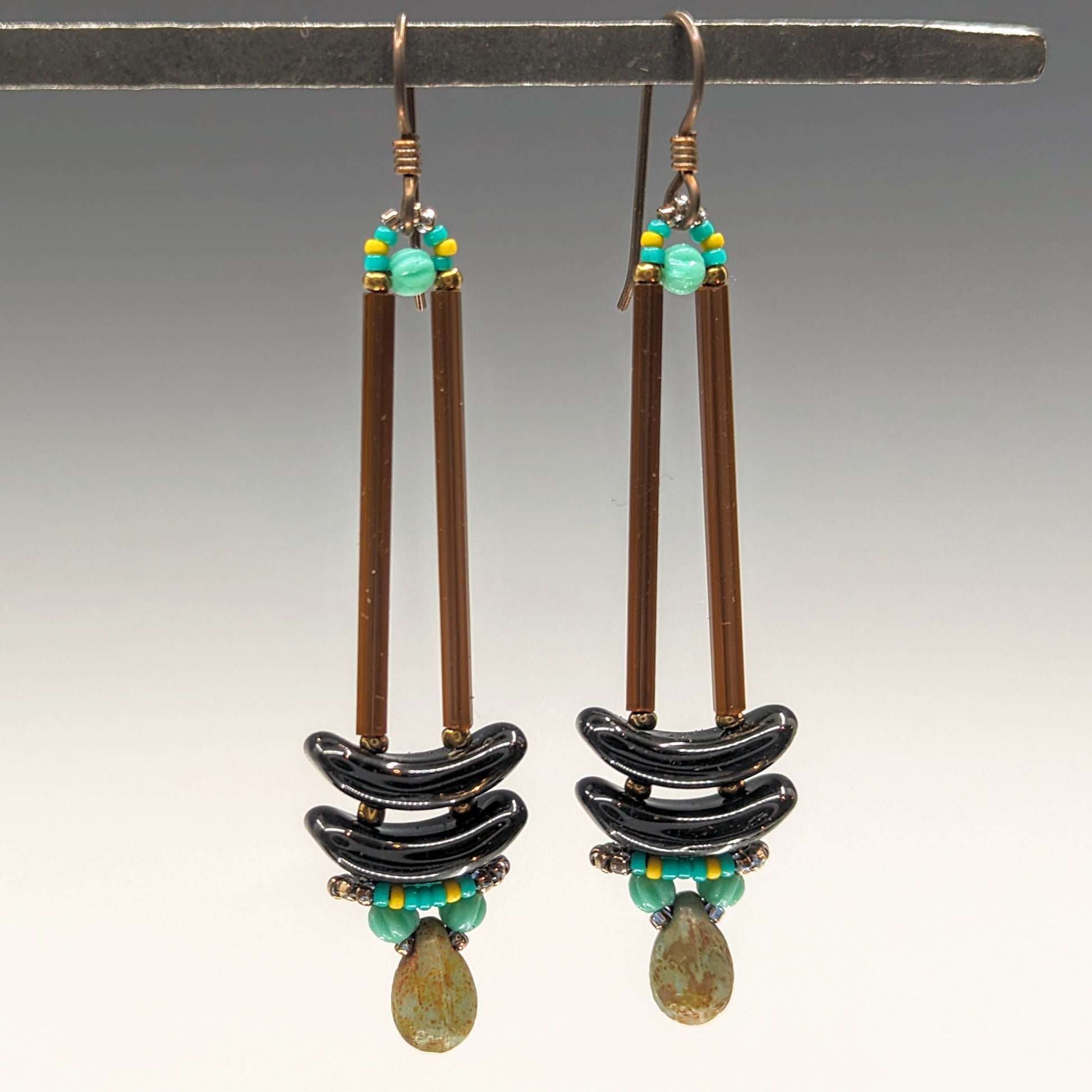 Earrings that look similar to a medicine dropper hang from  a black bar. There are two long, brown beads forming narrow vertical columns ending at the top of two black stacked arc shaped beads. At the bottom is a rustic turquoise drop shaped bead, surrounded by a belt of yellow and teal beads.