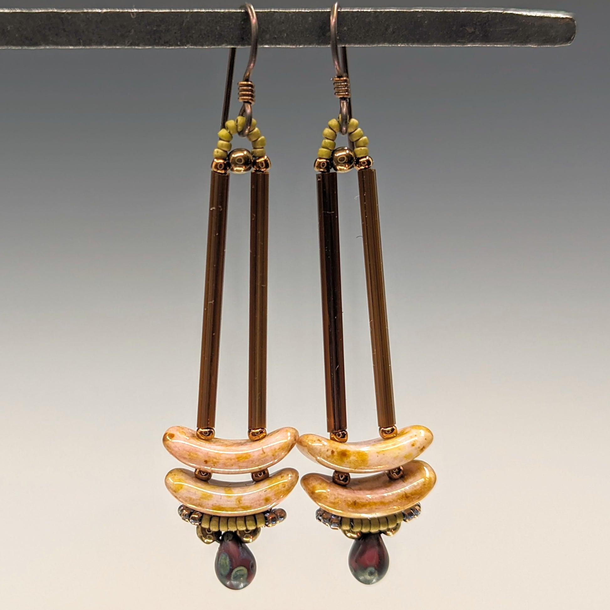 Earrings that look similar to a medicine dropper hang from  a black bar. There are two long, brown beads forming narrow vertical columns ending at the top of two speckled pink stacked arc shaped beads. At the bottom is a speckled red drop shaped bead, surrounded by a belt of gold seed beads.