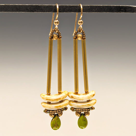 Earrings that look similar to a medicine dropper hang from a black bar. There are two long, yellow-gold beads forming narrow vertical columns ending at the top of two speckled cream stacked arc shaped beads. At the bottom is an avocado green drop shaped bead, surrounded by a belt of gold seed beads.