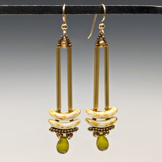 Earrings that look similar to a medicine dropper hang from a black bar. There are two long, yellow-gold beads forming narrow vertical columns ending at the top of two speckled cream stacked arc shaped beads. At the bottom is a matte transparent olive green drop shaped bead, surrounded by a belt of gold seed beads.