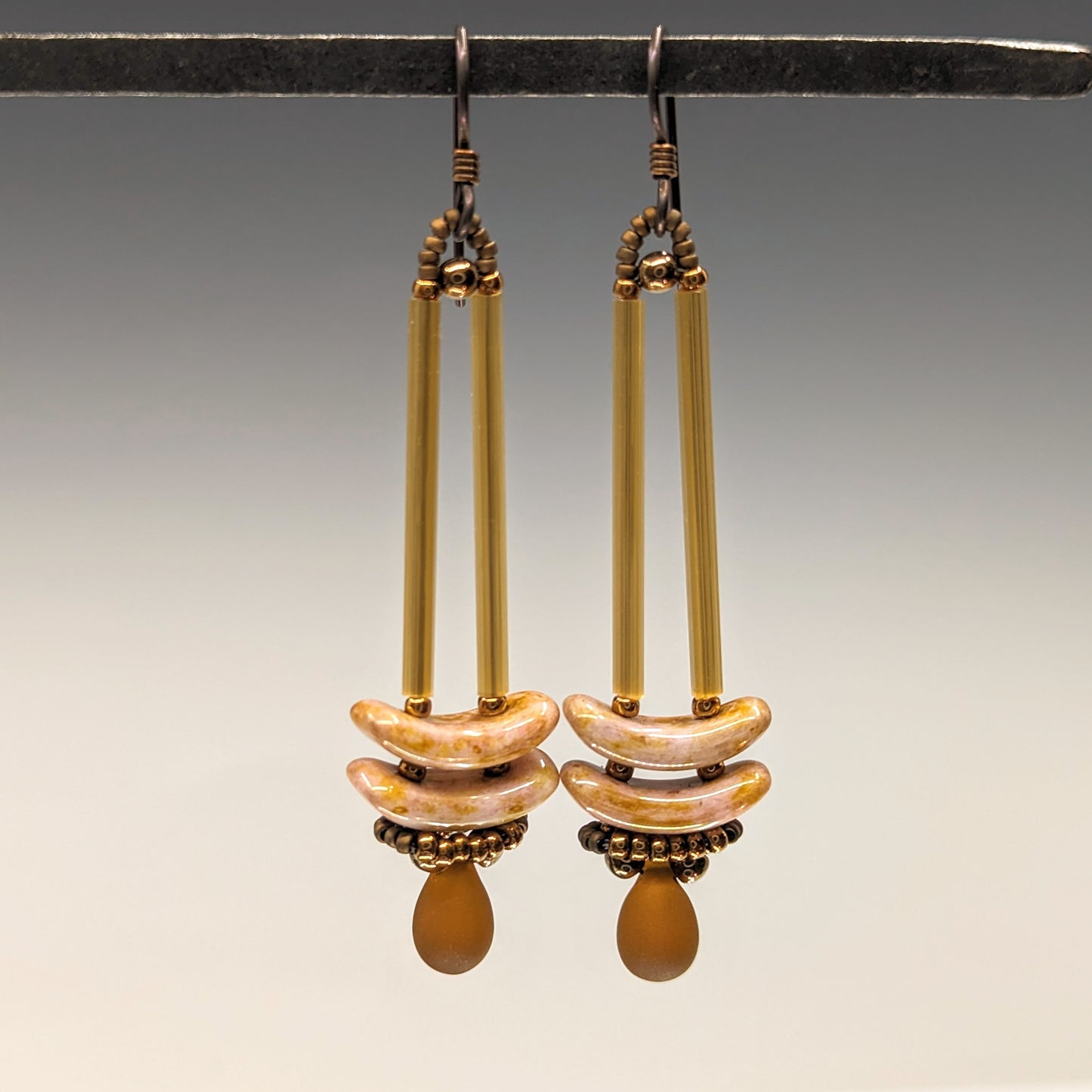 Earrings that look similar to a medicine dropper hang from  a black bar. There are two long, brown beads forming narrow vertical columns ending at the top of two speckled pink stacked arc shaped beads. At the bottom is a matte amber  drop shaped bead, surrounded by a belt of gold seed beads.