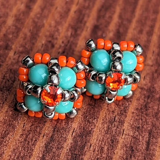 Square stud earrings resting on a reddish wood board. The earrings are formed from turquoise beads with an orange rhinestone. The rhinestone is held in place by an x-shape of silver seed beads. The outside of the earrings is outlined in orange seed beads. 