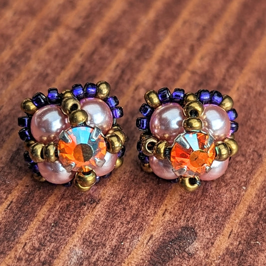 Square stud earrings formed from pearly pale pink beads with a golden-orange and rainbow flash rhinestone. The rhinestone is held in place by an x-shape of antique gold seed beads. The outside of the earrings is outlined in dark electric purple seed beads.