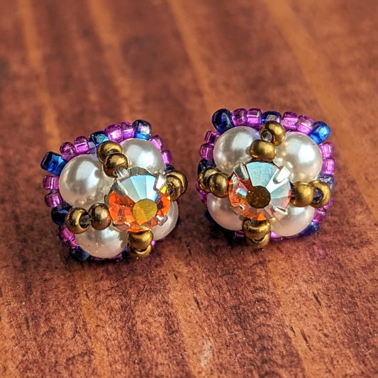 Square stud earrings resting on a reddish wood board. The earrings are formed from pearly white beads with a golden-orange and rainbow flash rhinestone. The rhinestone is held in place by an x-shape of antique gold seed beads. The outside of the earrings is outlined in grape purple and dark electric purple seed beads. 