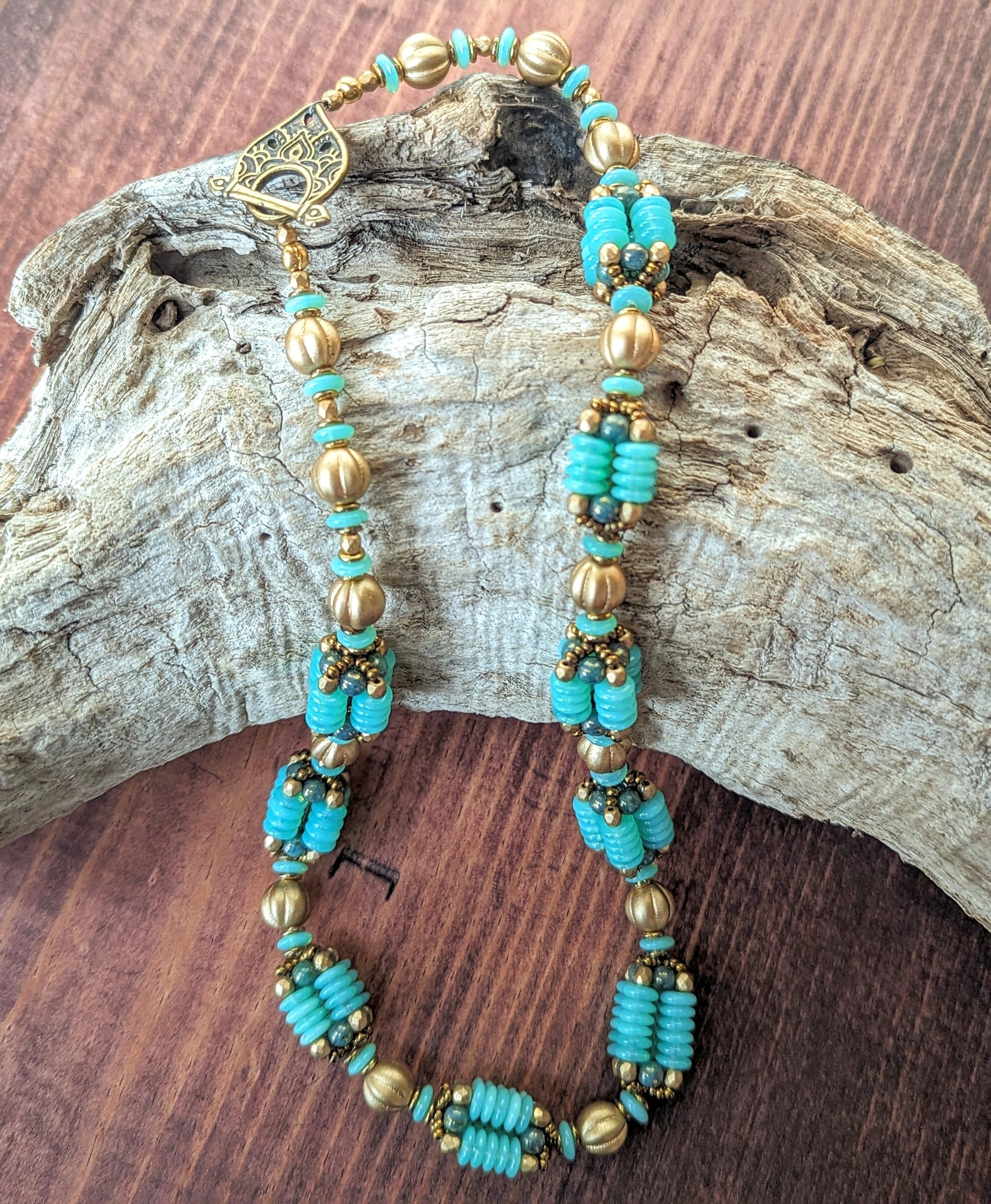 A light seafoam teal and gold necklace is draped across a piece of driftwood. The necklace has an alternating pattern of gold and seafoam beads, with nine handmade beaded beads with columns of light opalescent seafoam Czech glass discs with lustered teal accents. The necklace has a delicately patterned teardrop shaped brass toggle clasp.