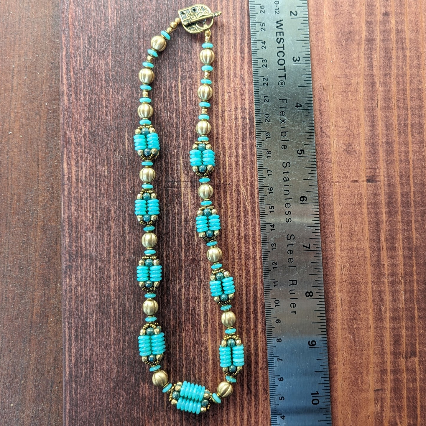 A light seafoam teal and gold necklace is laid out next to a ruler on a reddish wood board. The necklace has an alternating pattern of gold and seafoam beads, with nine handmade beaded beads with columns of light opalescent seafoam Czech glass discs with lustered teal accents. The necklace has a delicately patterned teardrop shaped brass toggle clasp.