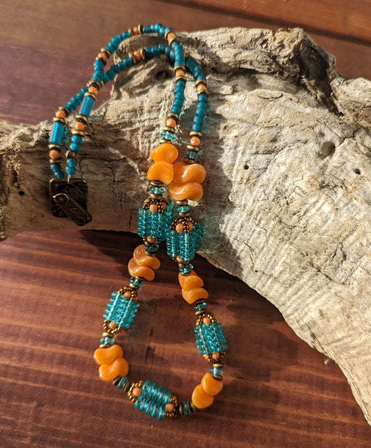 Lit by strong warm sunlight, a light orange and transparent teal necklace is draped across a light colored, curved piece of driftwood. The necklace has orange beads shaped like waves and teal beaded beads that are made up of four columns of teal discs. 