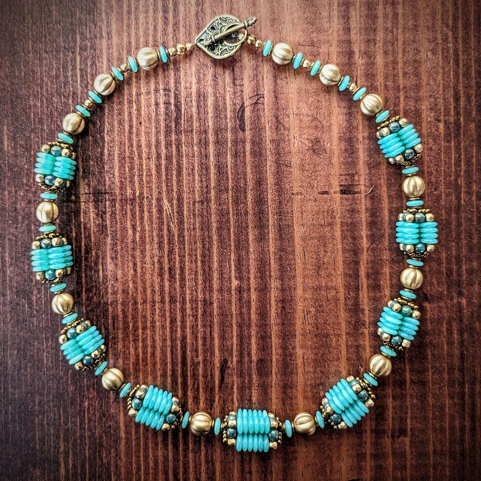 A light seafoam teal and gold necklace is laid out in a circle on a reddish wood board. The necklace has an alternating pattern of gold and seafoam beads, with nine handmade beaded beads with columns of light opalescent seafoam Czech glass discs with lustered teal accents. The necklace has a delicately patterned teardrop shaped brass toggle clasp.