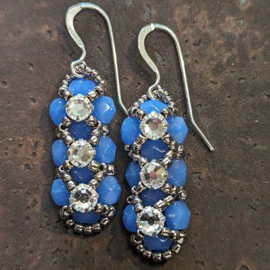 Silver and rich, lightly translucent medium blue earrings on a cork-like dark brown background. The earrings have a background of blue and there are silver seed beads in x-shape securing three clear rhinestones in a vertical row. The earrings have silver ear wires.