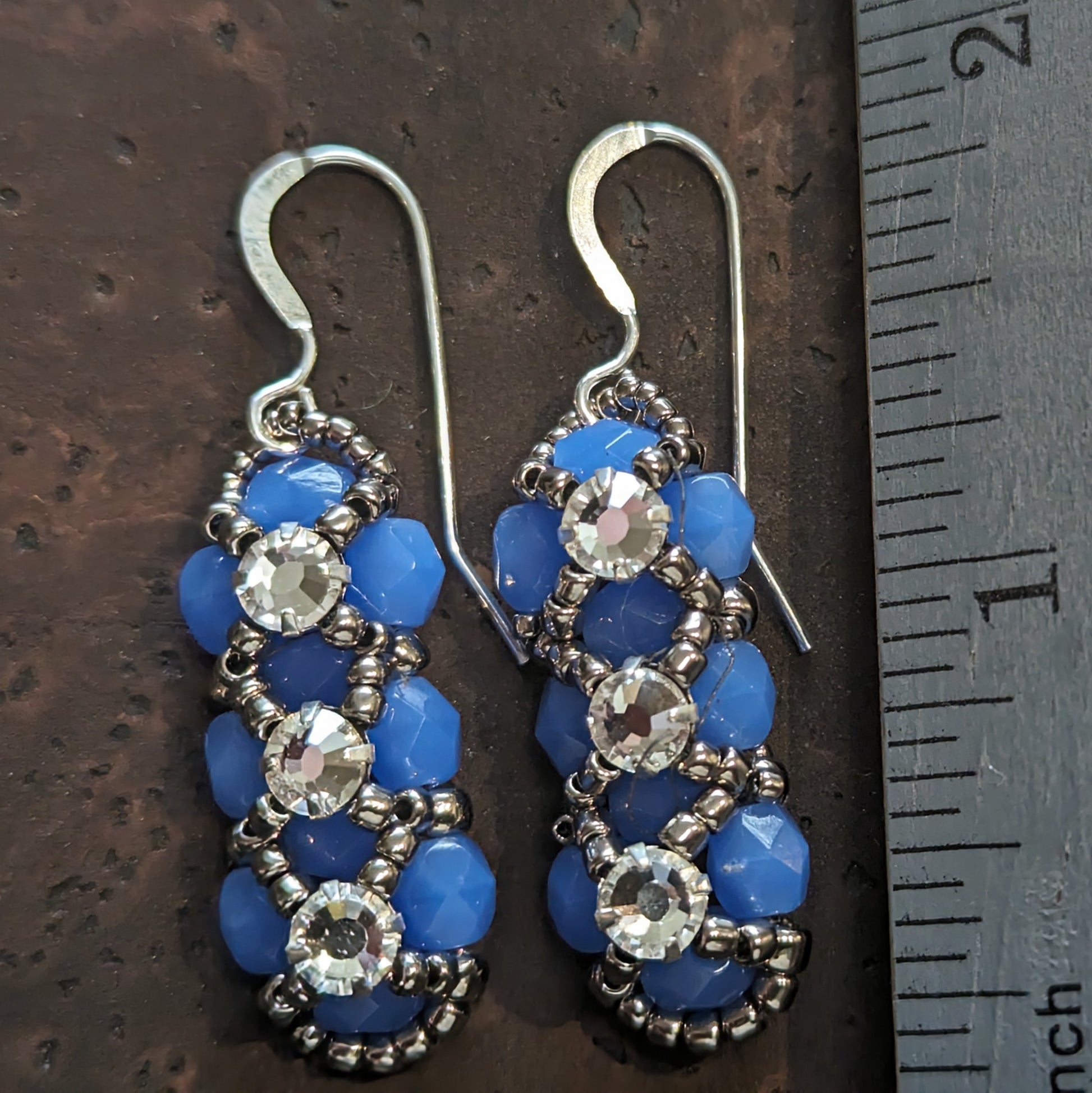 Silver and rich, lightly translucent medium blue earrings on a cork-like dark brown background next to a ruler.. The earrings have a background of blue and there are silver seed beads in x-shape securing three clear rhinestones in a vertical row. The earrings have silver ear wires.