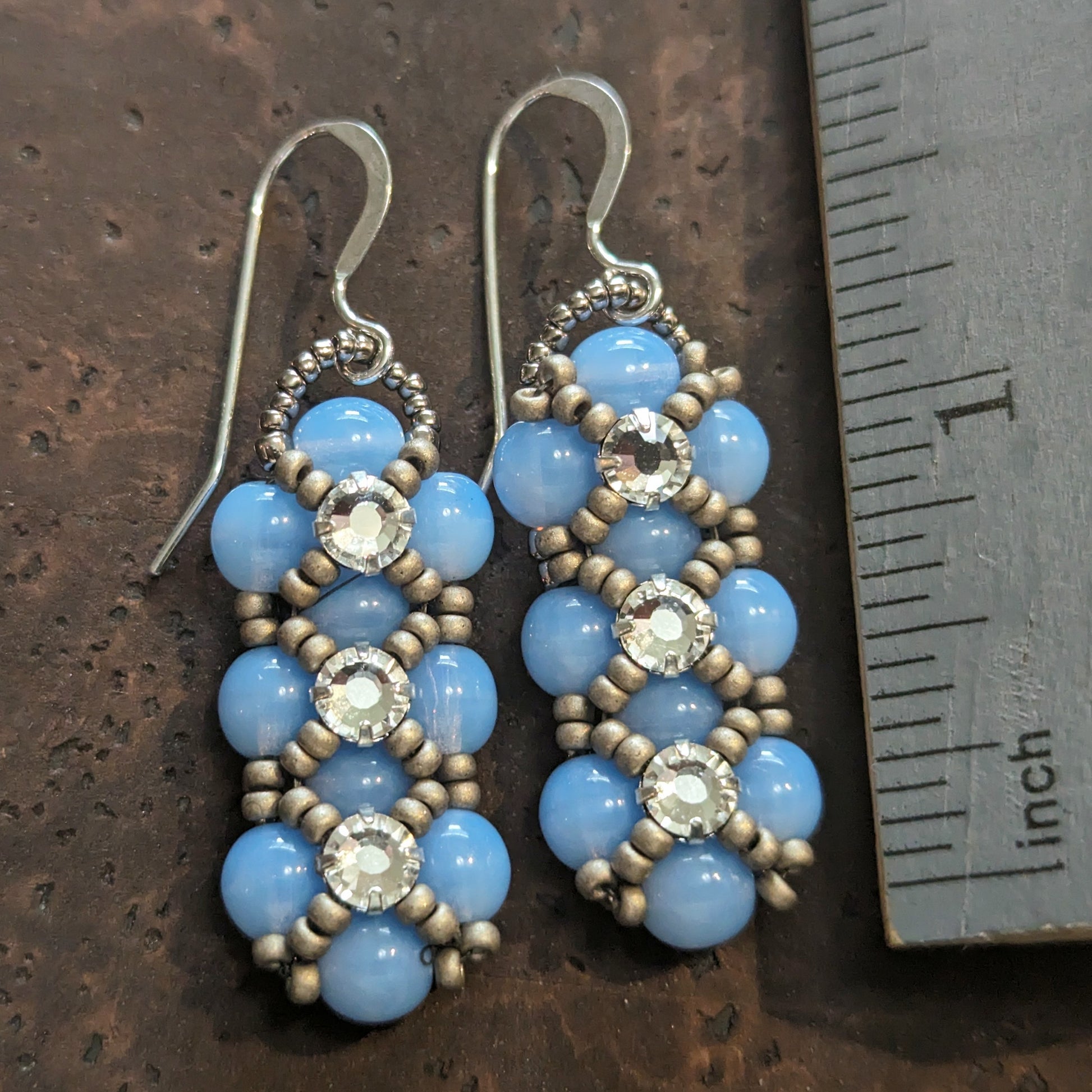 Silver and pale opal blue earrings on a cork-like dark brown background next to a ruler. The earrings have a background of light cloudy blue and there are matte silver seed beads in x-shape securing three clear rhinestones in a vertical row. The earrings have silver ear wires.