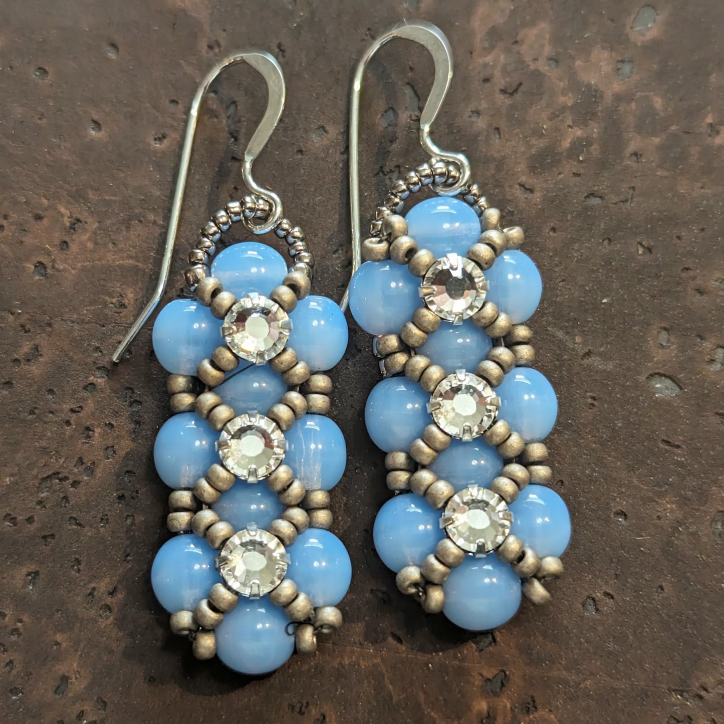Silver and pale opal blue earrings on a cork-like dark brown background. The earrings have a background of light cloudy blue and there are matte silver seed beads in x-shape securing three clear rhinestones in a vertical row. The earrings have silver ear wires.