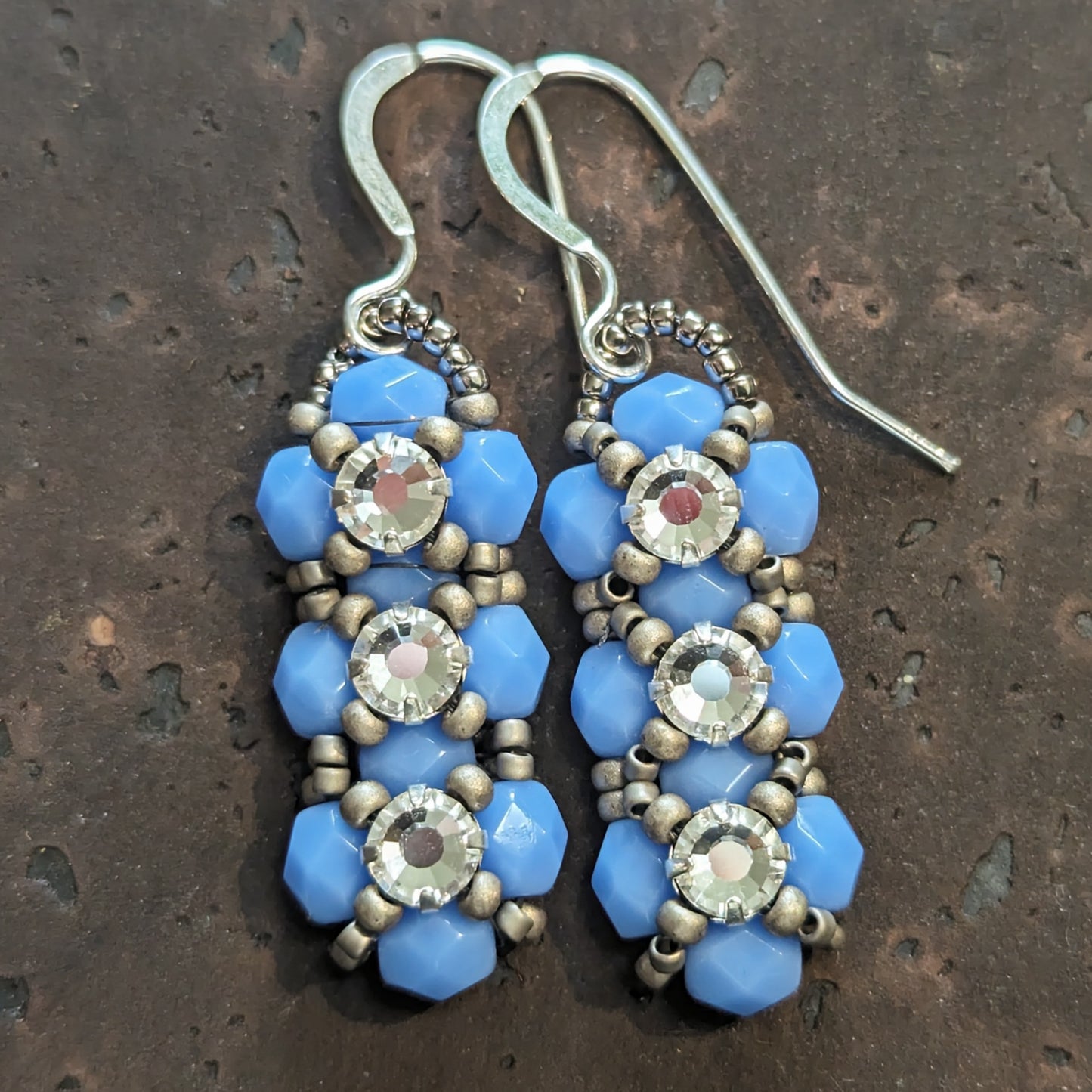 Silver and opaque pale periwinkle blue earrings on a cork-like dark brown background. The earrings have a background of light periwinkle blue and there are matte silver seed beads in x-shape securing three clear rhinestones in a vertical row. The earrings have silver ear wires.