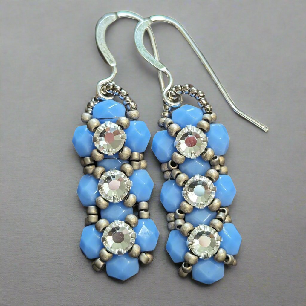 Silver and opaque pale periwinkle blue earrings on a cork-like dark brown background. The earrings have a background of light periwinkle blue and there are matte silver seed beads in x-shape securing three clear rhinestones in a vertical row. The earrings have silver ear wires.