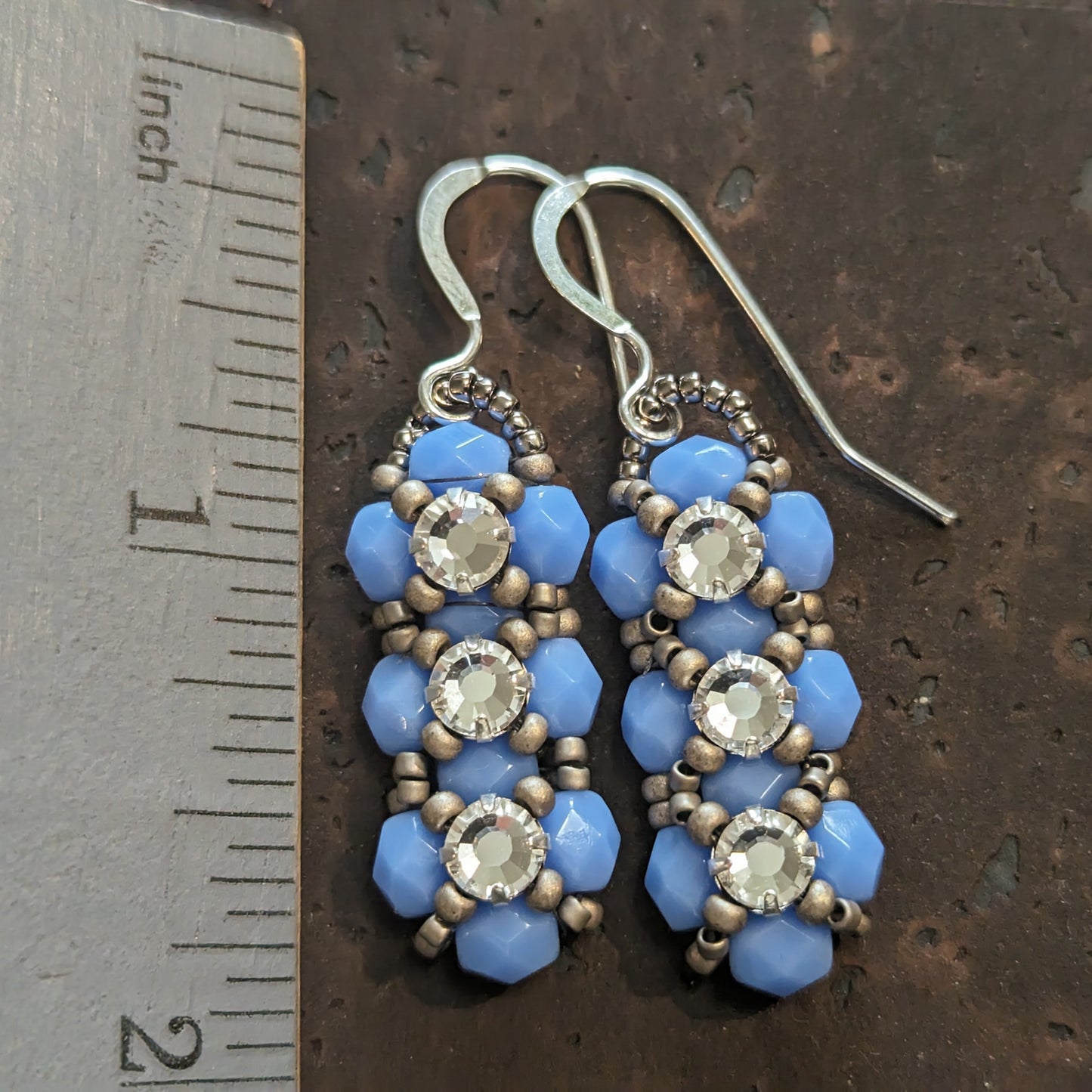 Silver and opaque pale periwinkle blue earrings on a cork-like dark brown background next to a ruler. The earrings have a background of light periwinkle blue and there are matte silver seed beads in x-shape securing three clear rhinestones in a vertical row. The earrings have silver ear wires.