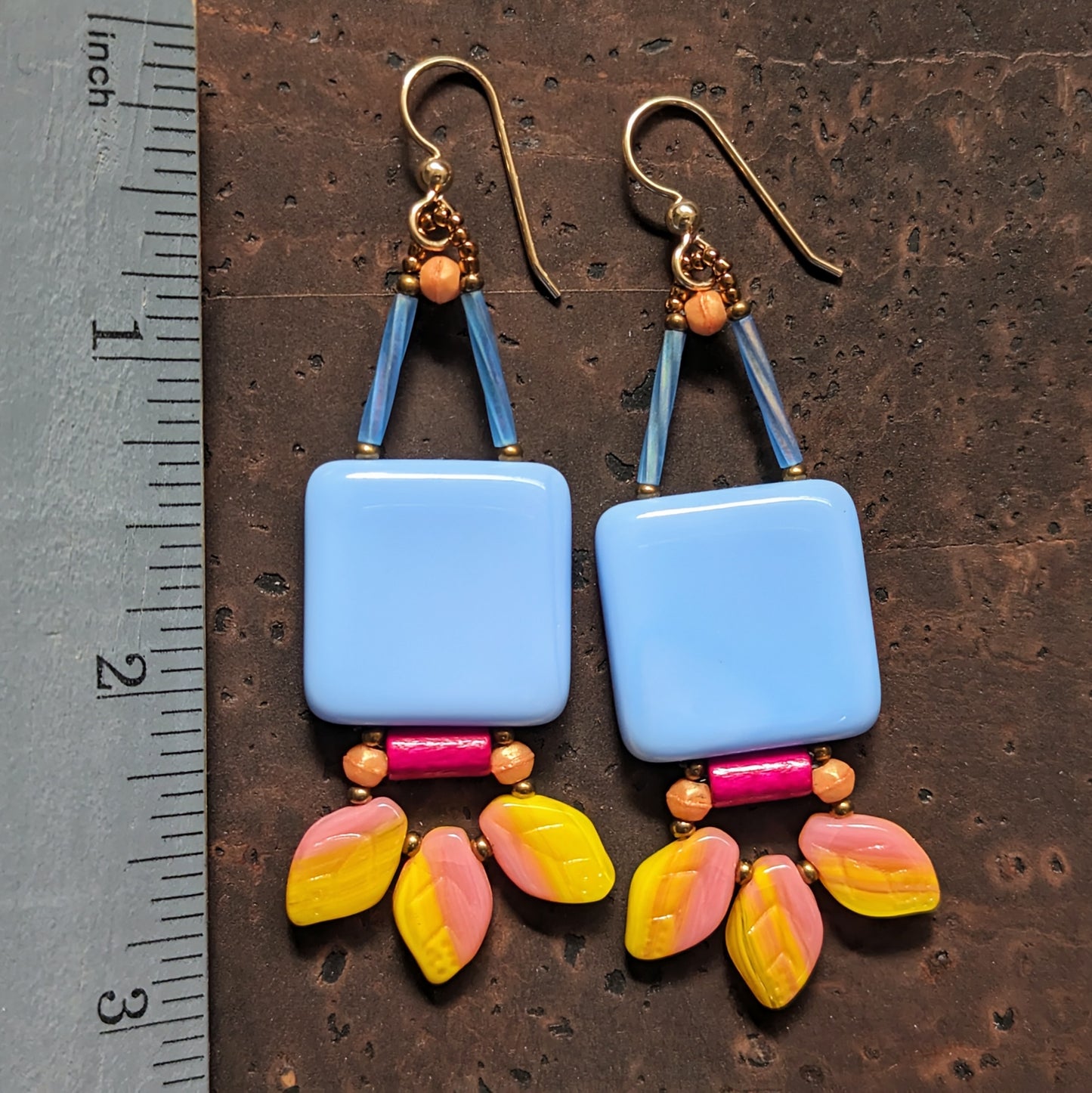 A pair of earrings lay on a dark brown cork background next to a ruler showing three inches as the length. The earrings are have a large light blueish square suspended from gold wires. Below the square is a horizontal bright pink tube with apricot beads on either side. At the bottom is a swag of three leaf shaped beads made from variegated yellow and pastel pink glass.