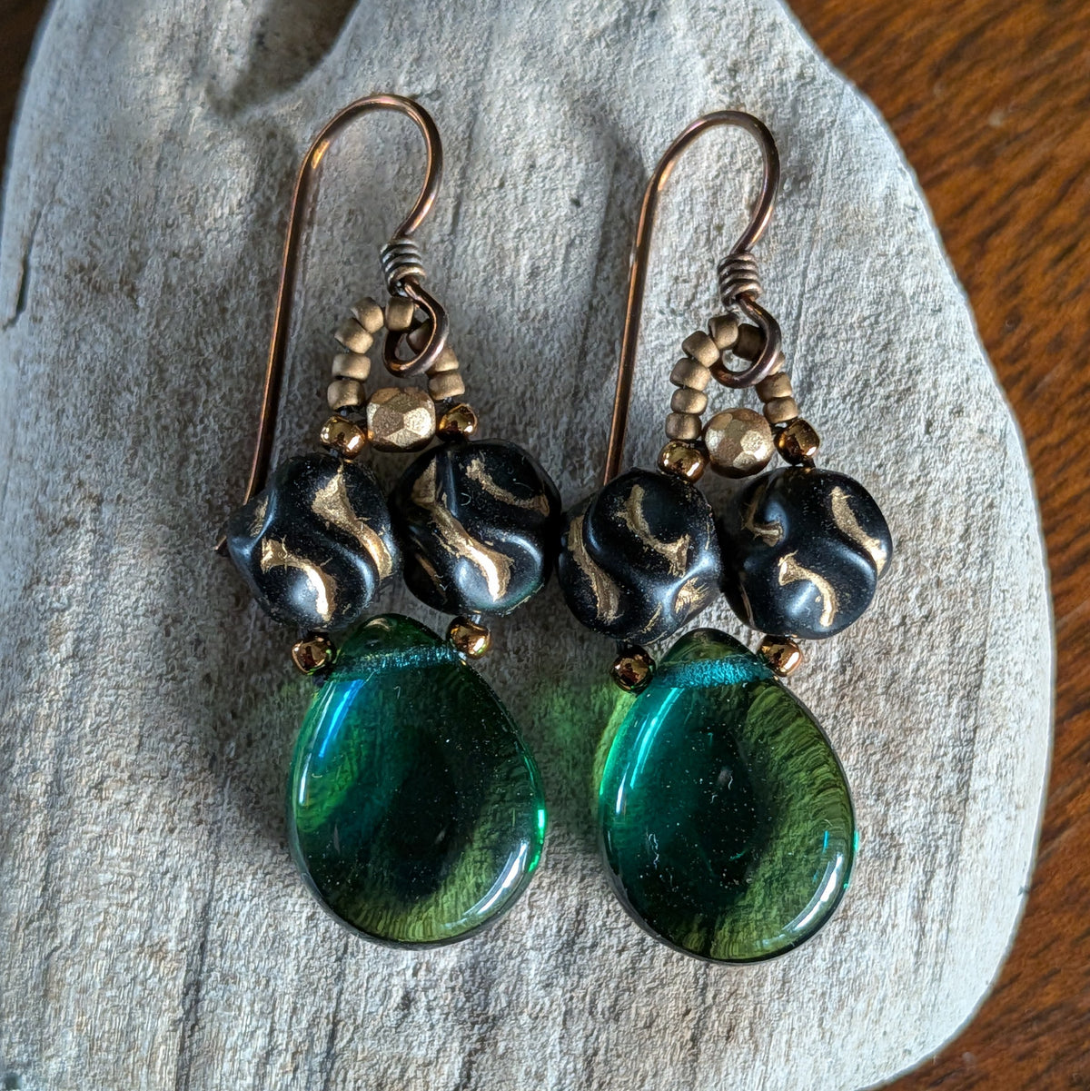 Pair of earrings with green teardrop beads and black beads with a gold pattern on a wooden surface.