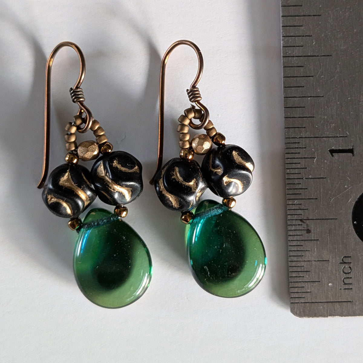 Pair of earrings with green teardrop-shaped beads and gold accents on a wooden surface with a ruler for scale.