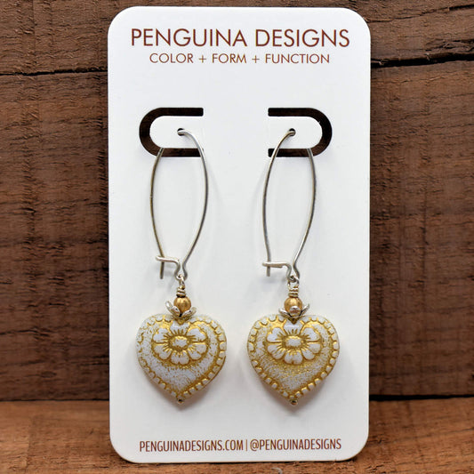 A pair of earrings on a white card rest against a wood background. The earrings have long silver  oval wires that latch and white hearts at the bottom with gold accents. 